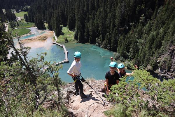 Three people wearing helmets and harnesses are engaged in a climbing or rappelling activity on a rocky cliff, overlooking a stunning turquoise lake surrounded by dense pine forests